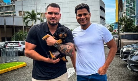 Two men smiling at camera in urban parking lot, one holding a small black and tan dog