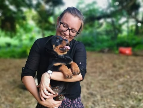 Young woman wearing glasses holds a small black and tan puppy in a garden setting