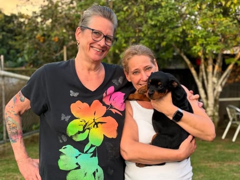 Two adults smiling outdoors in a backyard, one wearing a colorful flower graphic t-shirt and holding a black dog
