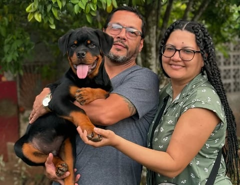 A man and woman holding a Rottweiler dog outdoors with green foliage in the background