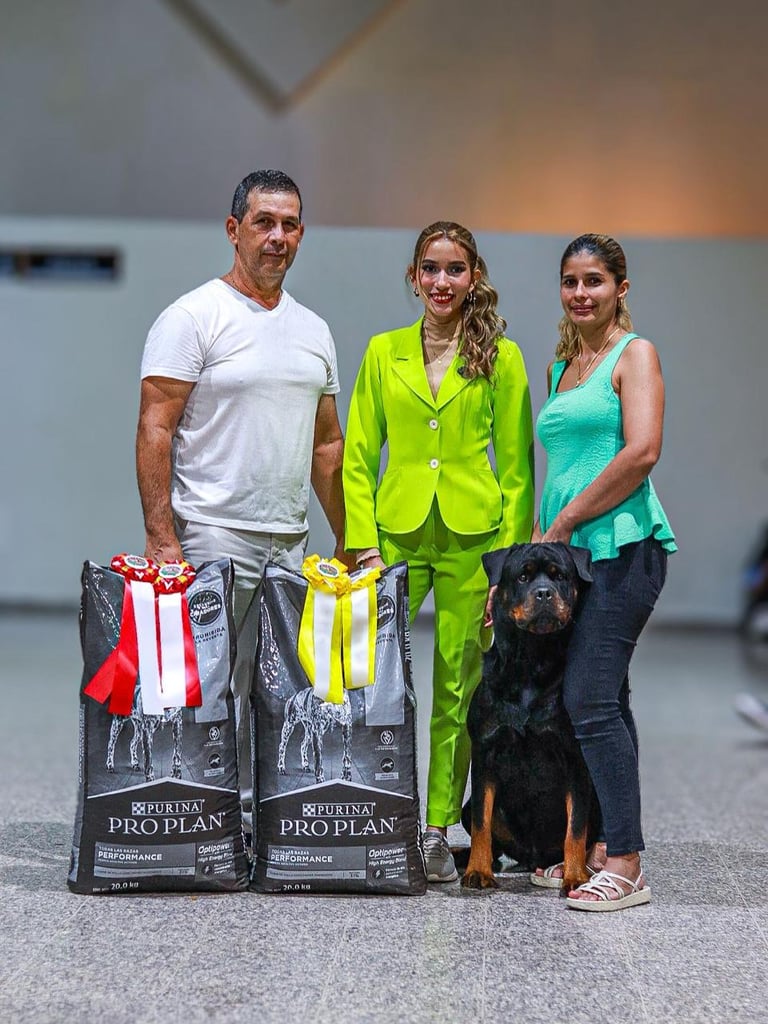 Three people and a black dog standing with Purina Pro Plan dog food bags and award ribbons at an indoor event