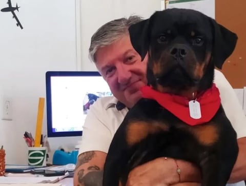 A man holding a black Rottweiler dog wearing a red bandana, smiling at the camera in an office setting